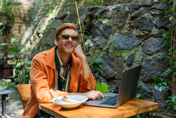 happy redhead freelancer in sunglasses using laptop near prepared lunch and cocktail on table - Photo, image