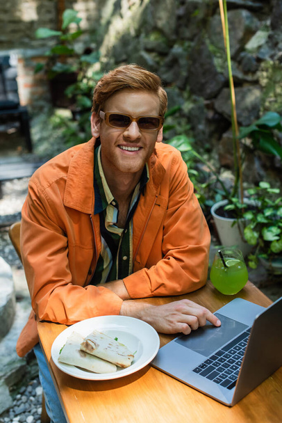 cheerful redhead freelancer in sunglasses using laptop near prepared lunch and cocktail on table - Фото, зображення