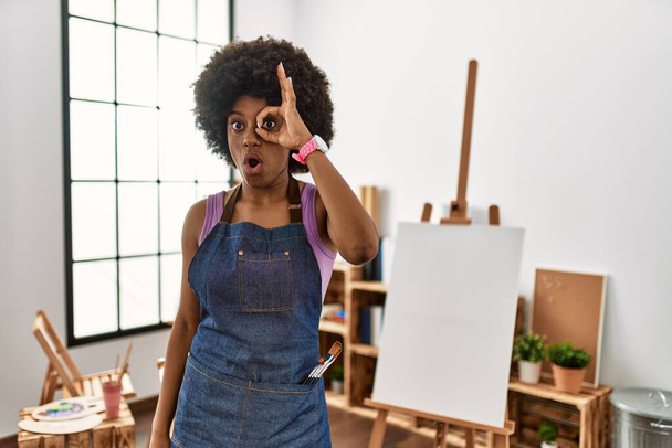 Mujer afroamericana joven con pelo afro en el estudio de arte haciendo buen gesto conmocionado con la cara sorprendida, los ojos mirando a través de los dedos. expresión incrédula.  - Foto, imagen
