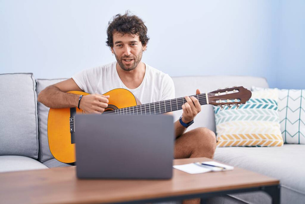 Молодий іспаномовний чоловік має онлайн класичний гітарний клас, сидячи на дивані вдома
 - Фото, зображення