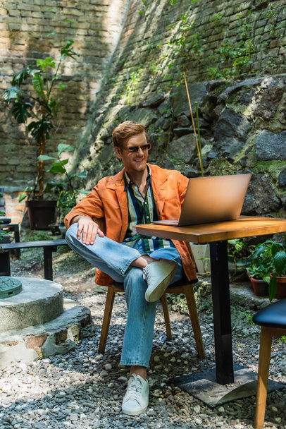 happy redhead man in sunglasses working remotely on laptop while sitting in outdoor terrace - Foto, Imagen