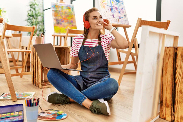 Jovem mulher sorrindo confiante usando laptop e fones de ouvido no estúdio de arte - Foto, Imagem