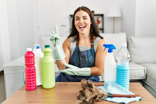 Young brunette woman wearing cleaner apron and gloves cleaning at home smiling with happy face winking at the camera doing victory sign. number two.  - Photo, Image