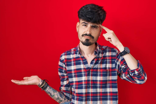 赤い背景の上に立って髭を生やした若いヒスパニック系の男は混乱し、コピースペースを示し、額に指を指すオープンヤシに悩まされました。考えてみてください.  - 写真・画像