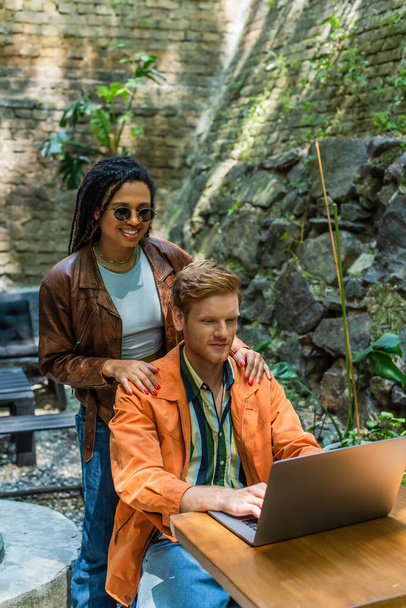 happy african american woman touching shoulders of redhead man using laptop in outdoor terrace  - Photo, image