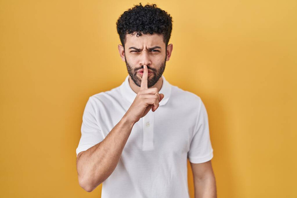 Hombre árabe de pie sobre fondo amarillo pidiendo silencio con el dedo en los labios. silencio y concepto secreto.  - Foto, imagen