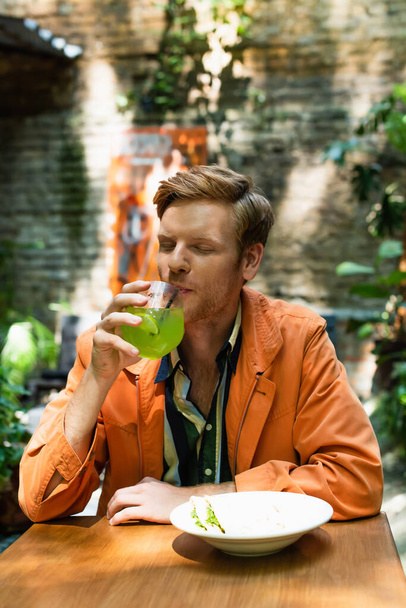 redhead man drinking cocktail near prepared lunch in outdoor cafe terrace - Photo, image