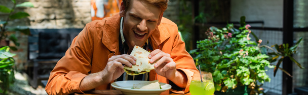веселий рудий чоловік їсть квазаділла біля келиха з коктейлем на відкритій терасі, банер
 - Фото, зображення