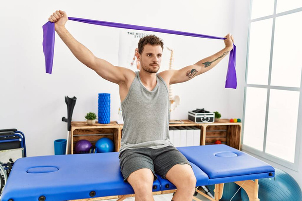 Jonge Spaanse man stretching arm met behulp van elastische band zitten op massage boord in kliniek - Foto, afbeelding