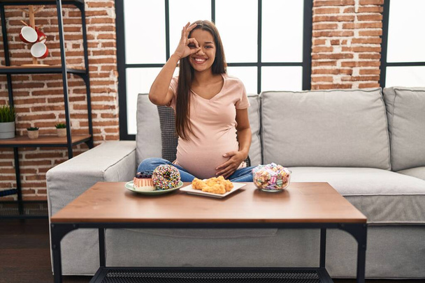 Νεαρή έγκυος γυναίκα τρώει γλυκά στο σπίτι χαμογελώντας ευτυχισμένη κάνει εντάξει σημάδι με το χέρι στο μάτι κοιτάζοντας μέσα από τα δάχτυλα  - Φωτογραφία, εικόνα