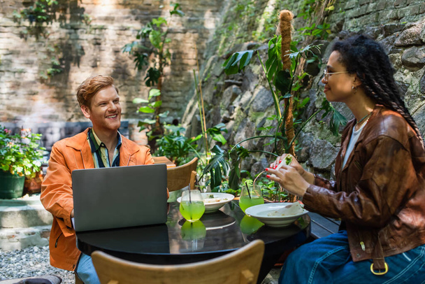 african american woman holding quesadilla near happy freelancer friend using laptop in outdoor terrace - Photo, Image