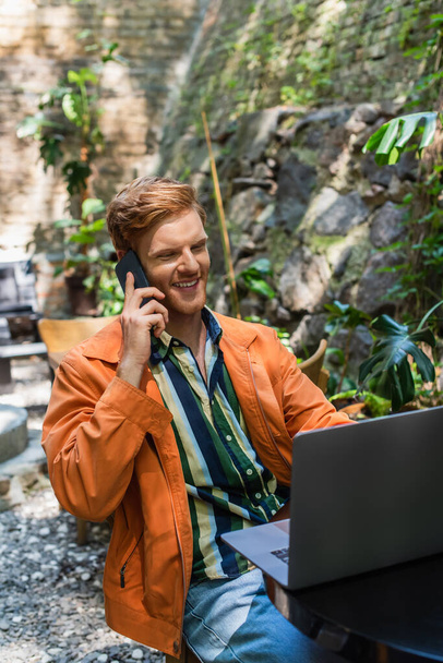 cheerful redhead man talking on smartphone while using laptop in cafe terrace - Фото, зображення