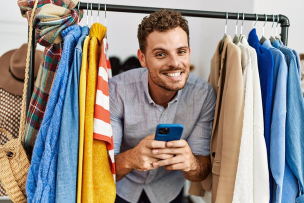 Mladý hispánský zákazník muž usměvavý šťastný objevuje přes oblečení pomocí smartphone v obchodě s oblečením. - Fotografie, Obrázek