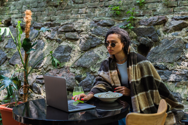 african american freelancer in sunglasses covered in blanket using laptop and having lunch in cafe on terrace - 写真・画像