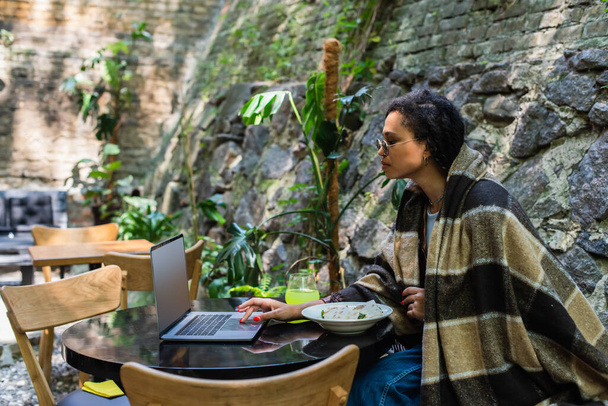 african american freelancer covered in blanket using laptop and having lunch in cafe on terrace - Foto, afbeelding