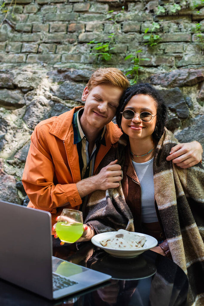 happy redhead man hugging african american friend covered in blanket while having lunch in cafe near laptop - Photo, image