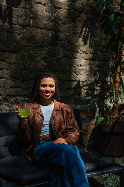 happy african american woman in sunglasses holding glass with green cocktail and sitting outside - Foto, Imagen