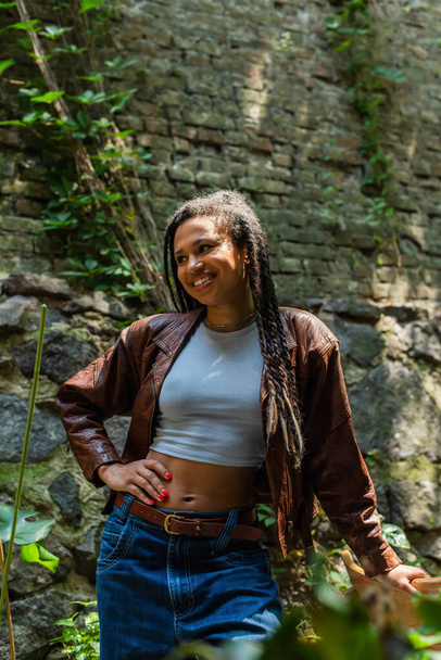 щаслива афроамериканка в коричневій шкіряній куртці, що стоїть з рукою на стегні
  - Фото, зображення