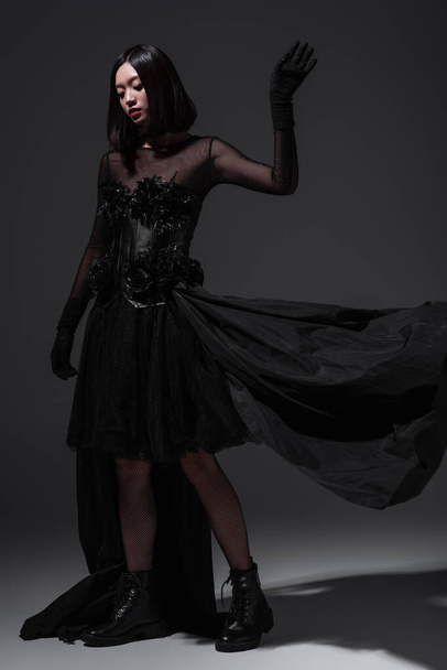 full length of young asian woman in black gothic dress and gloves posing on dark grey - Foto, Imagem