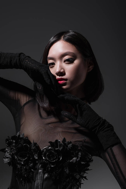 brunette asian woman in black gothic outfit and gloves posing isolated on dark grey - Photo, Image