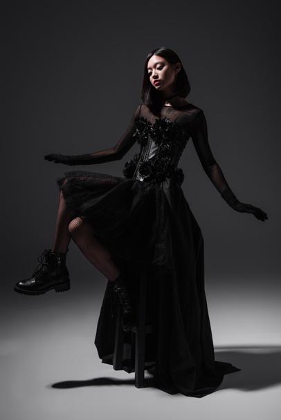 полная длина азиатской молодой женщины в черно-готическом платье и перчатках, сидящих на высоком стуле на темно-сером - Фото, изображение