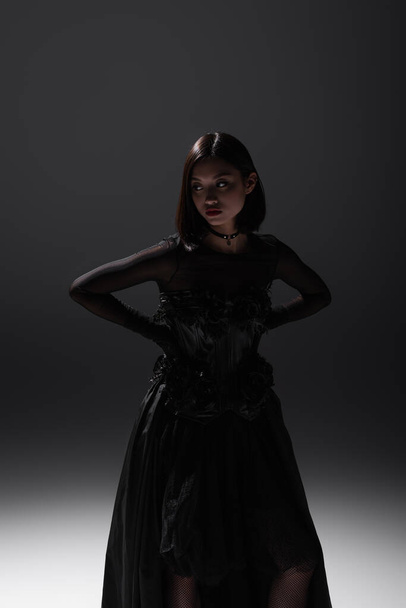 brunette asian model in black gothic dress posing with hands on hips on dark grey - Fotó, kép