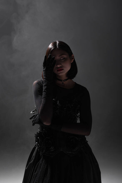 brunette asian model with closed eyes posing in black gloves and gothic outfit on dark grey with smoke - Photo, image