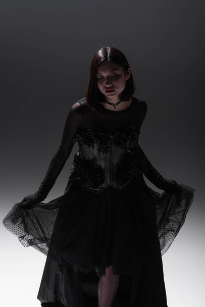 asian model in black gothic dress holding lace skirt while posing on grey - Foto, Imagem