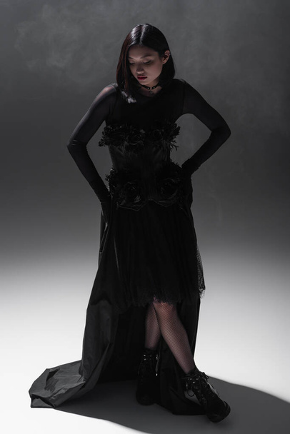 full length of asian young woman in black gothic dress and gloves posing on dark grey with smoke - Valokuva, kuva