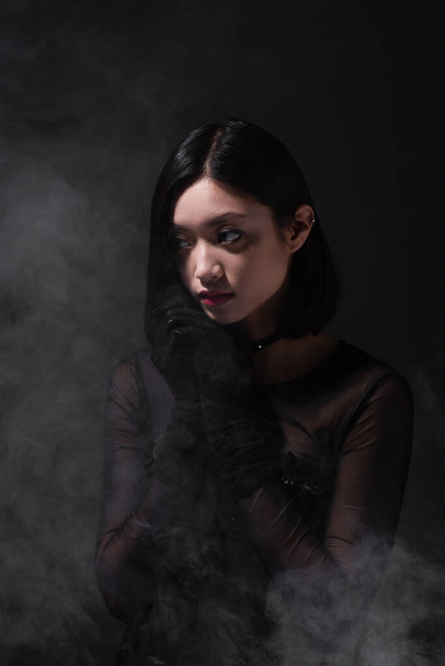 portrait of young asian woman in gothic outfit and gloves looking away on grey with smoke - Fotoğraf, Görsel