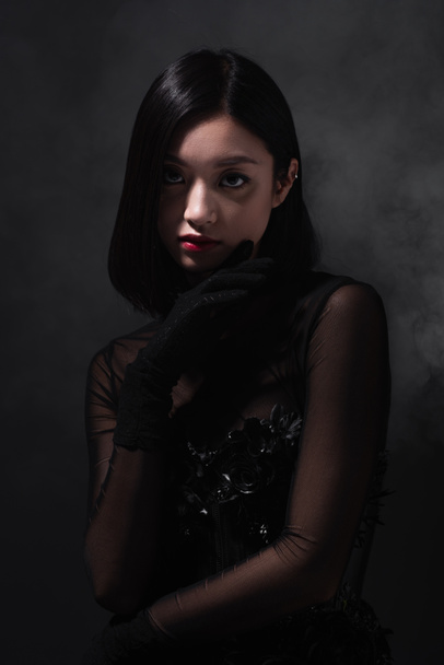 portrait of young asian woman in gothic outfit and gloves looking at camera on grey with smoke - Foto, Imagen