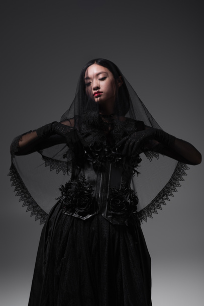 молодая азиатская модель в готическом наряде с черной вуалью, позирующая изолированно на сером - Фото, изображение