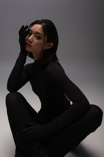 stylish and young asian model in black outfit with gloves sitting on grey   - Foto, immagini