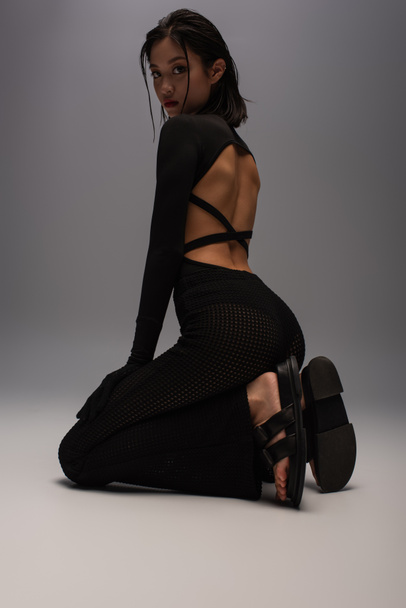 full length of brunette asian woman in total black and stylish outfit sitting on knees on grey background  - Fotó, kép