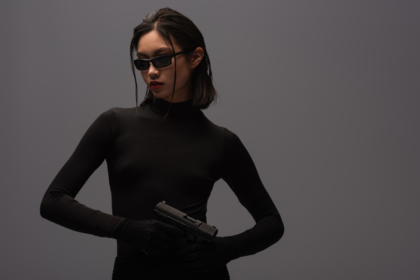 озброєна азіатська жінка в загальному чорному вбранні і стильні сонцезахисні окуляри тримають пістолет ізольовано на сірому
 - Фото, зображення