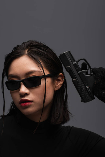 portrait of dangerous asian woman in total black outfit and stylish sunglasses holding gun isolated on grey - Foto, Imagem