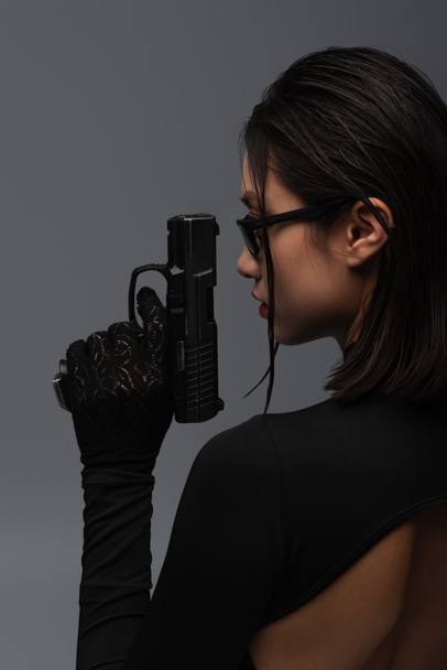 side view of dangerous asian woman in total black outfit and stylish sunglasses holding gun isolated on grey - Fotó, kép