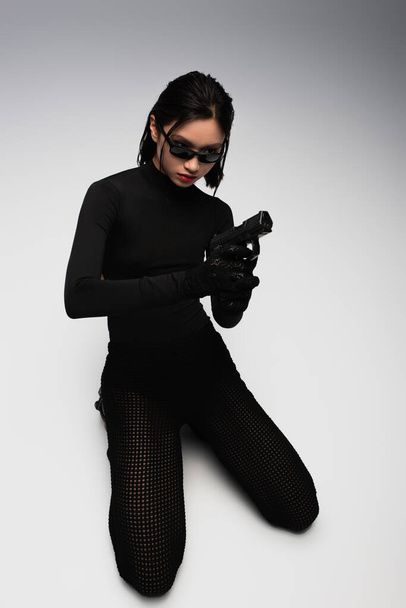 опасная азиатка в черном костюме и стильных солнцезащитных очках, держащая пистолет на белом - Фото, изображение