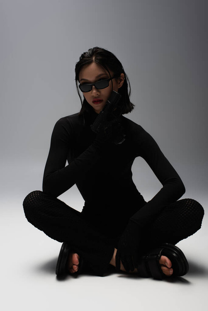 азиатка в черном костюме и стильных солнцезащитных очках, держащая пистолет, сидя на сером  - Фото, изображение