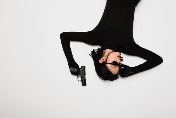 top view of brunette asian woman in black outfit adjusting stylish sunglasses and holding gun while lying on white - Zdjęcie, obraz