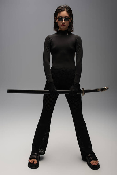 полная длина азиатская женщина в черном наряде и стильные солнцезащитные очки держа катана меч на сером - Фото, изображение