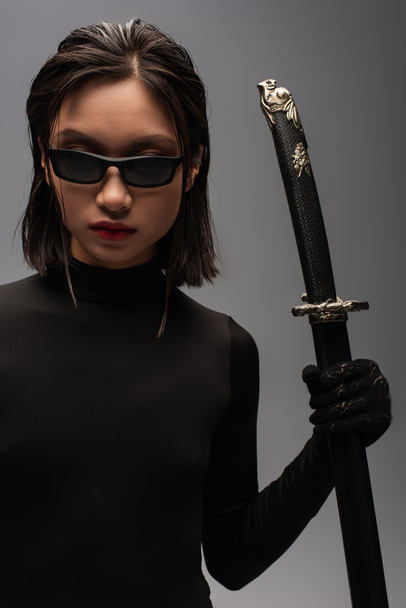 brunette asian woman in black clothes and stylish sunglasses holding katana sword in scabbard isolated on grey - Photo, Image