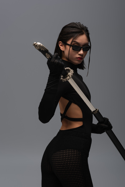 dangerous asian woman in black outfit and stylish sunglasses pulling out katana from scabbard isolated on grey - Fotó, kép