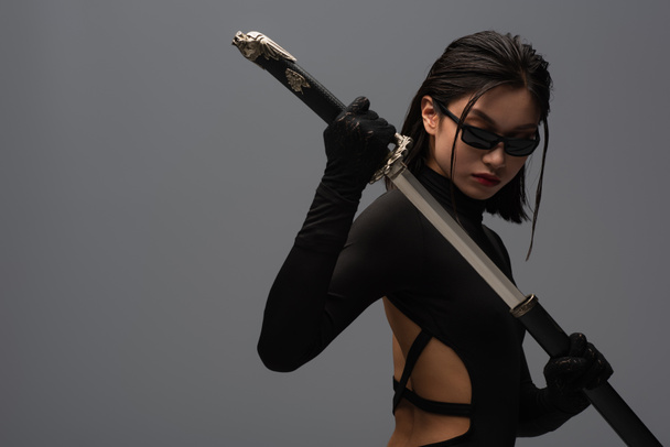 brunette asian woman in black clothes and sunglasses pulling out katana from scabbard isolated on grey - Foto, Bild