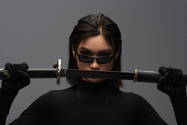 front view of asian woman in black outfit and stylish sunglasses pulling out katana from scabbard isolated on grey - Fotó, kép