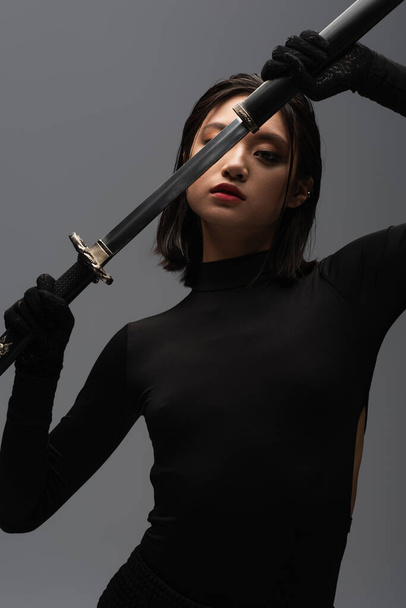 brunette asian woman in black outfit pulling out katana from scabbard isolated on grey - Fotó, kép
