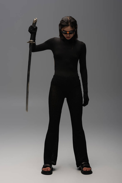 full length of brunette asian woman in sunglasses and black outfit holding katana on grey - Photo, image