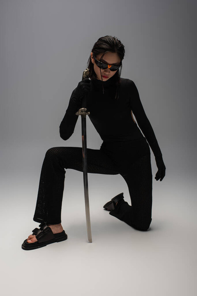 full length of dangerous asian woman in sunglasses and black clothes holding katana while standing on knee on grey - Fotó, kép