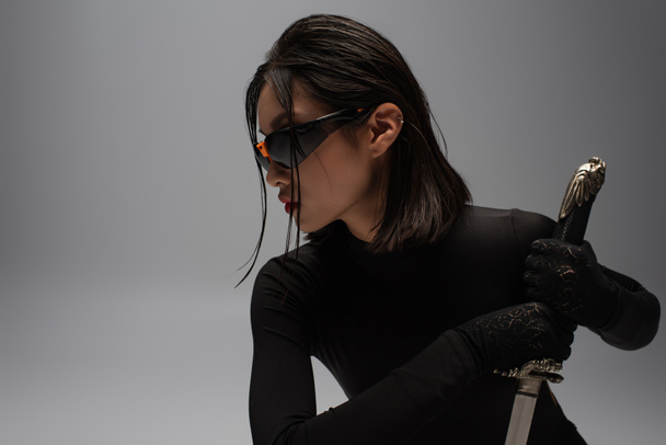side view of dangerous asian woman in sunglasses and black outfit holding katana isolated on grey - Foto, Imagen