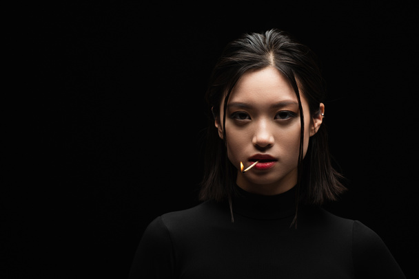 portrait of pretty asian woman with burning match in mouth looking at camera isolated on black - Photo, Image